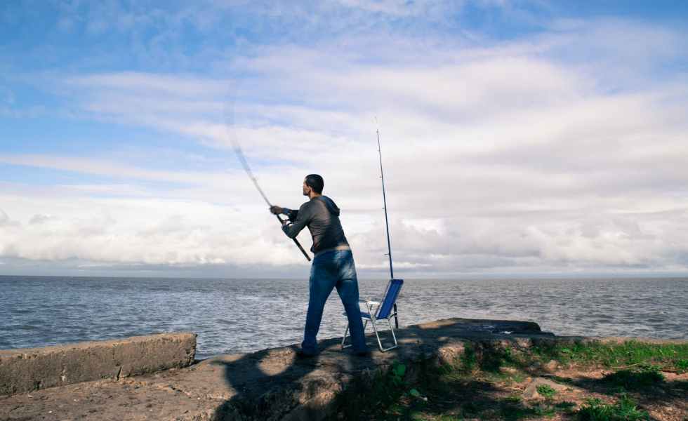 man in blue shirt and blue denim jeans holding fishing rod standing on brown rock during